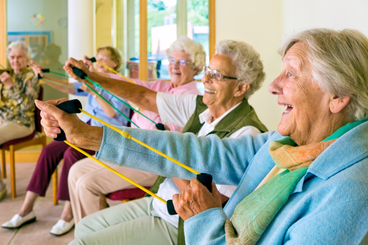 Large group of happy enthusiastic elderly ladies exercising in a gym sitting in chairs doing stretching exercises with rubber bands.